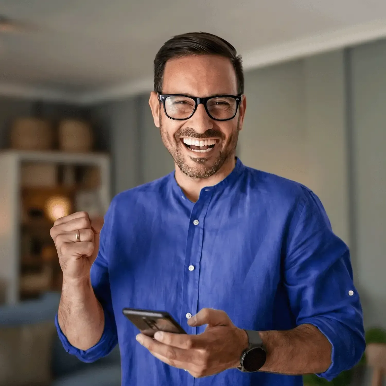 Homem sorridente de &oacute;culos e camisa azul comemorando com o celular na m&atilde;o.