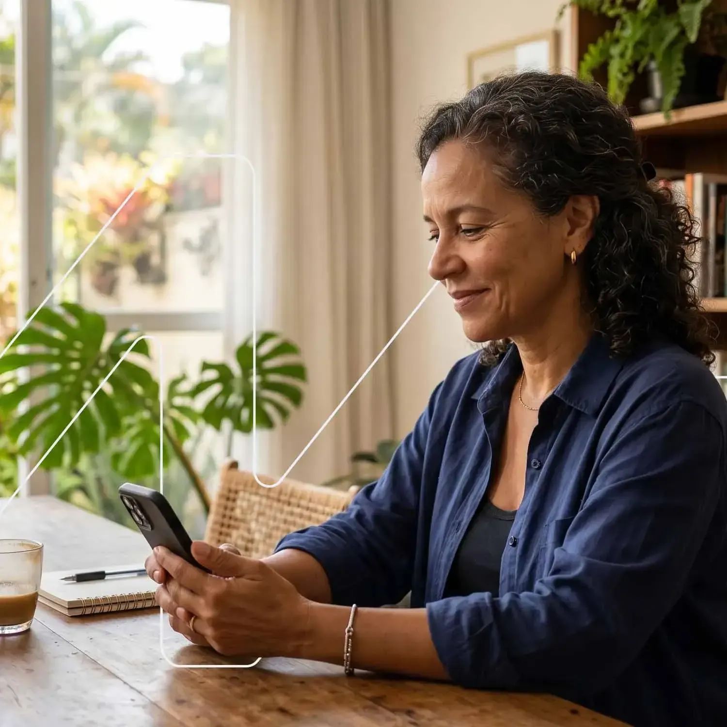 Mulher madura sorrindo ao usar o celular em uma mesa de madeira, com plantas ao fundo e grafismos em linhas brancas na imagem.