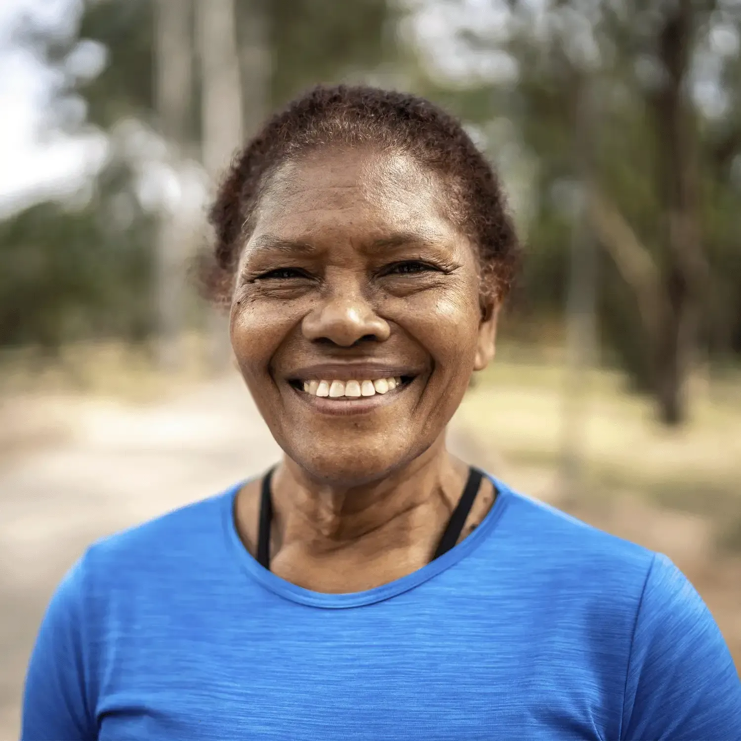 Mulher sorrindo, vestindo camiseta azul, com fundo de natureza desfocado.