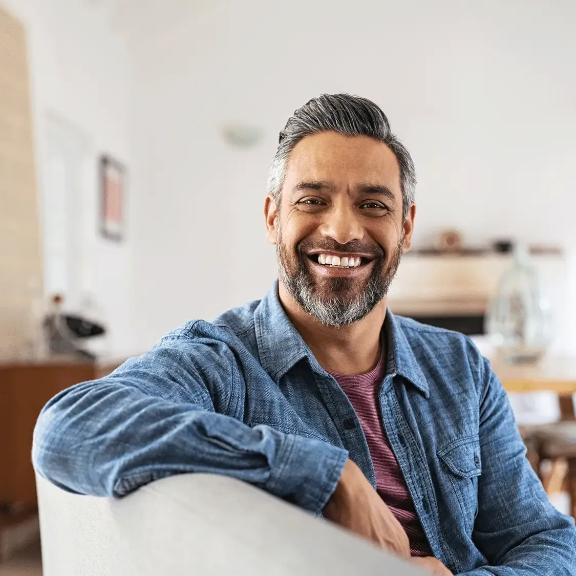 Homem de meia-idade grisalho sorrindo no sof&aacute; de casa, com ambiente ao fundo levemente desfocado.