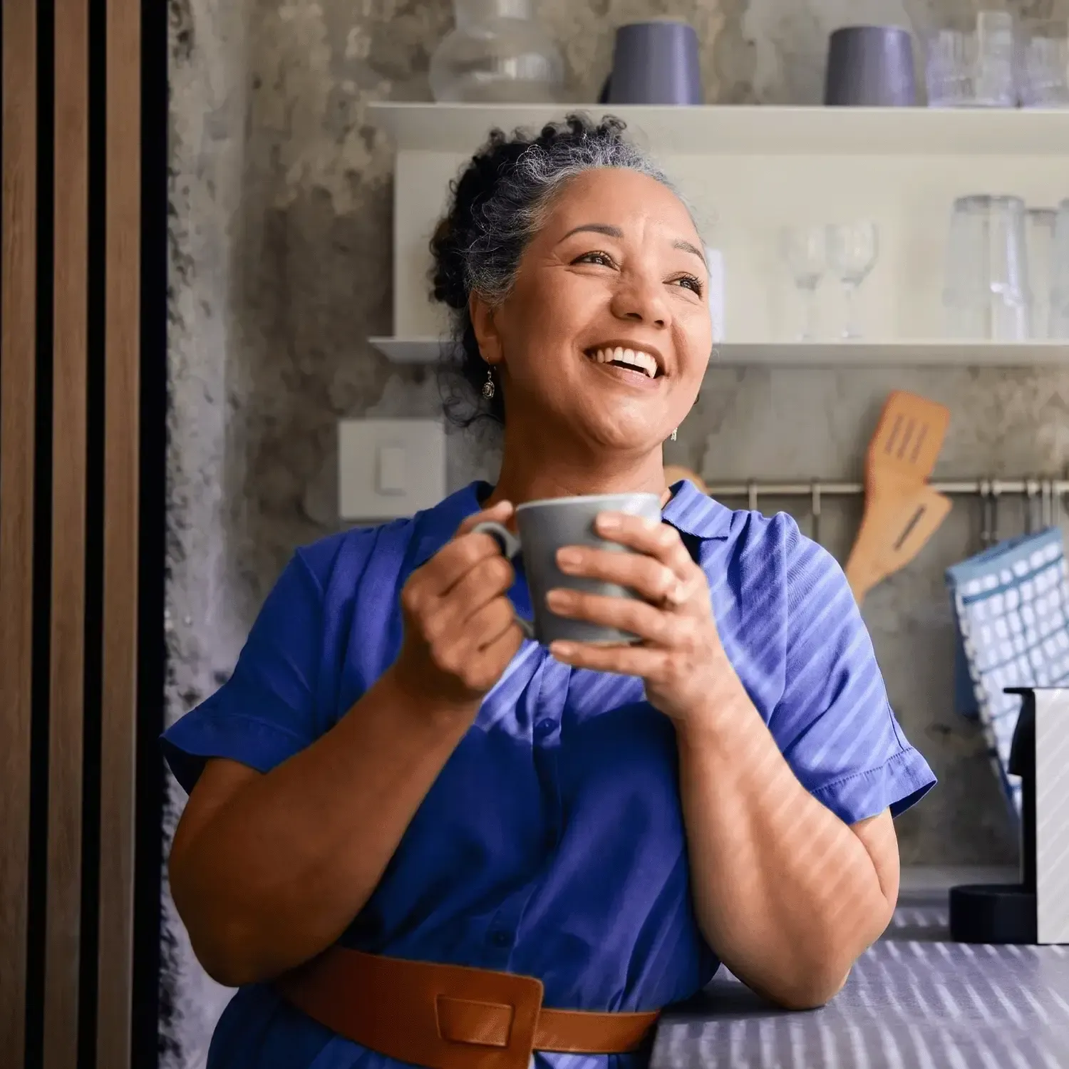 Mulher madura e sorridente segurando uma caneca de caf&eacute; no balc&atilde;o da cozinha.