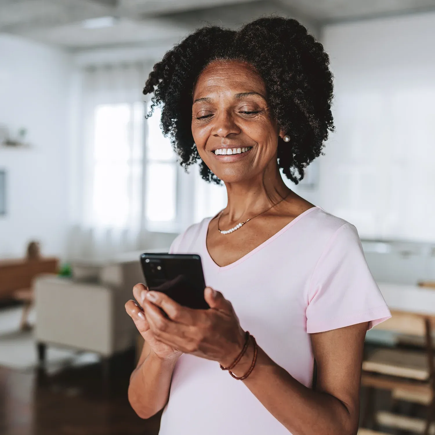 Mulher negra madura sorrindo ao usar o smartphone na sala de casa.