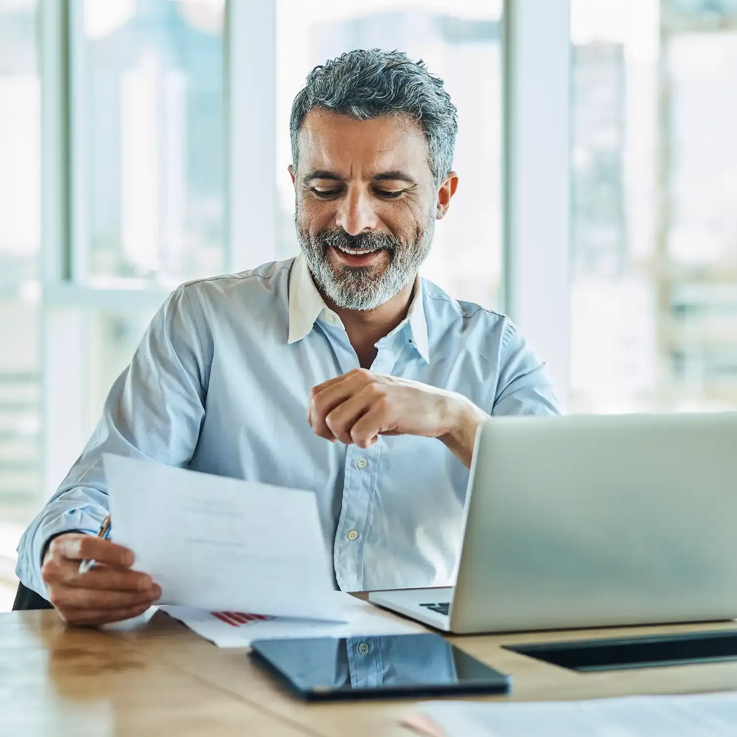 Homem sorridente lendo um relat&oacute;rio financeiro ao lado de seu notebook no escrit&oacute;rio.