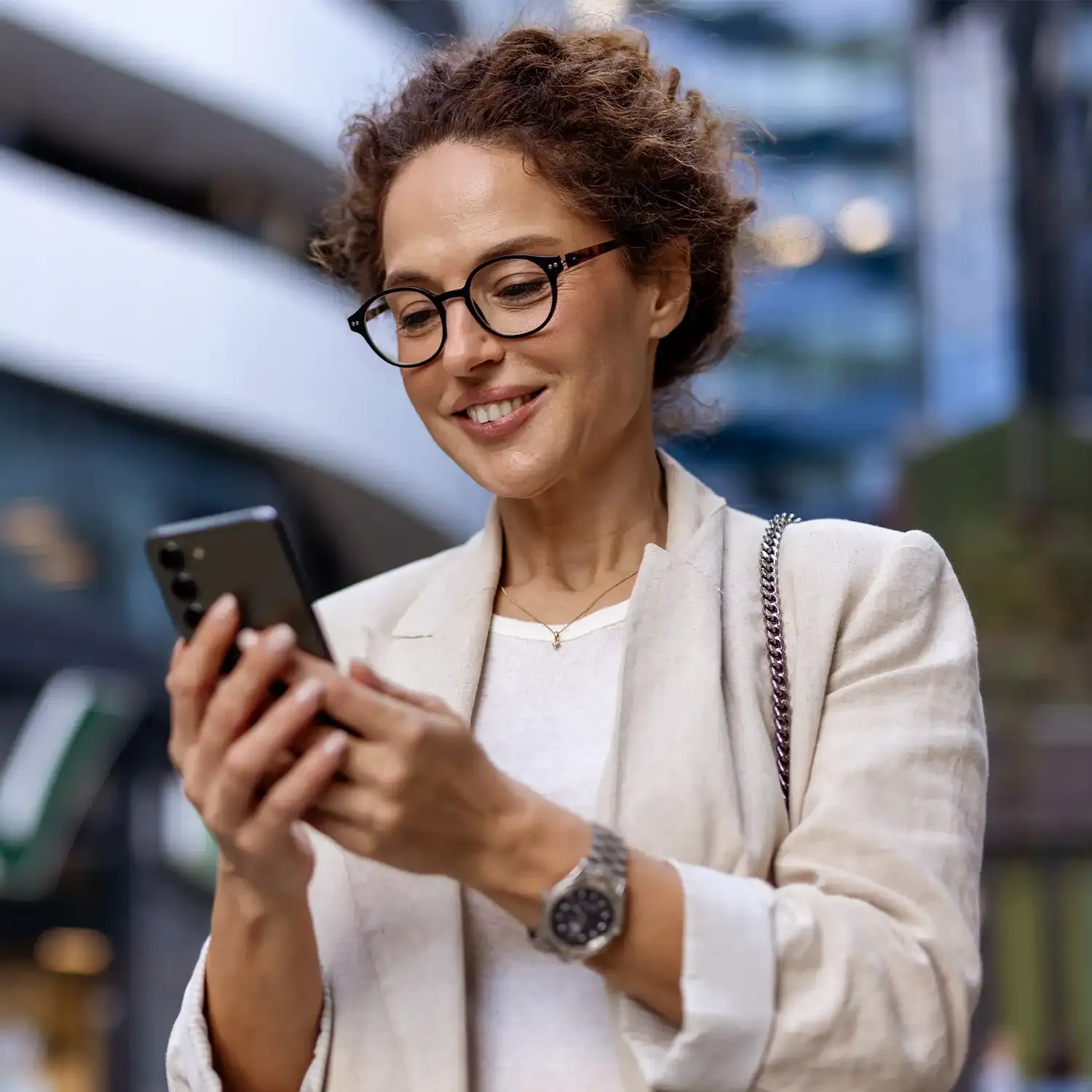 Mulher elegante de &oacute;culos sorrindo ao olhar o celular caminhando na cidade.