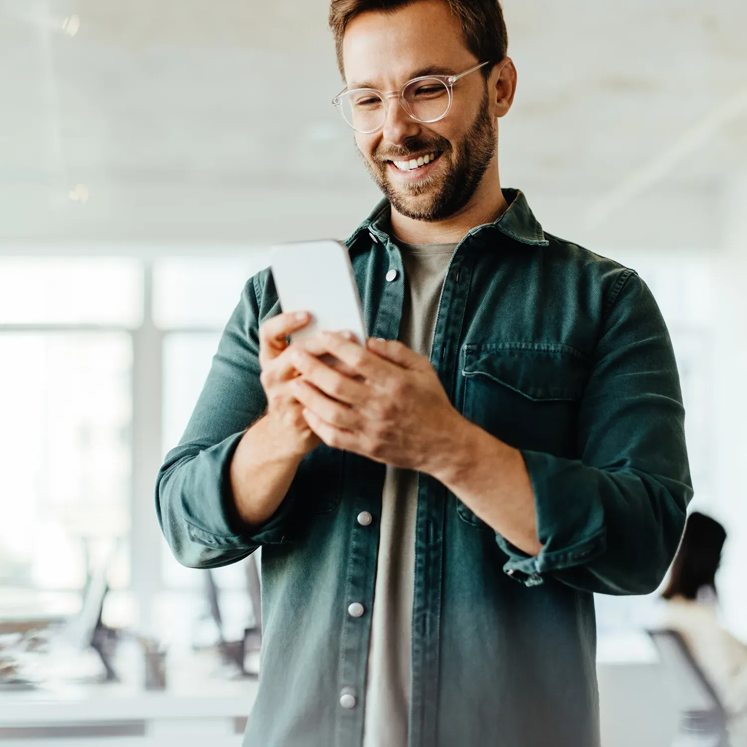 Homem de &oacute;culos sorrindo para a tela do celular em um ambiente de trabalho.