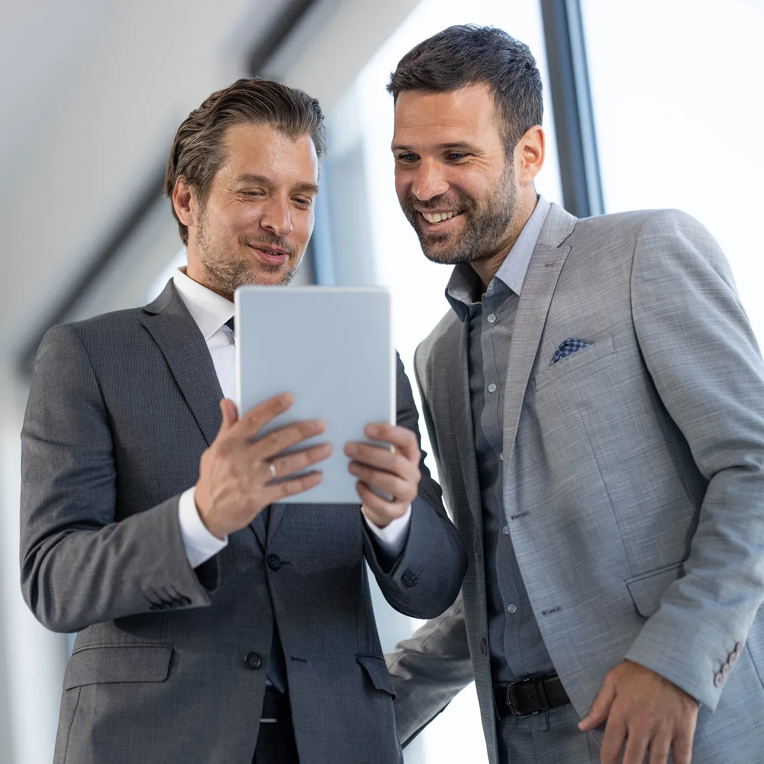 Dois homens de terno sorrindo e olhando para um tablet em ambiente corporativo.